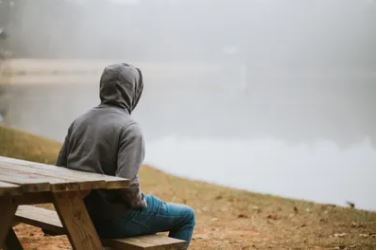 Photo of a person sitting at a wooden outdoor table on the shore of a body of water obscured by fog or mist. The person is wearing a hooded shirt and is facing away from the viewer, toward the water.