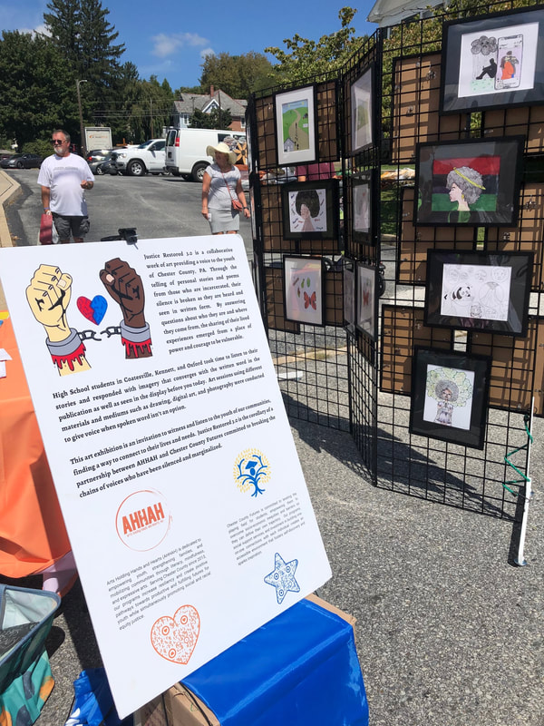 A poster describing the Justice Restored Traveling Art Exhibit stands in front of a folding wire screen displaying some of the artwork in frames in a paved outdoor area.